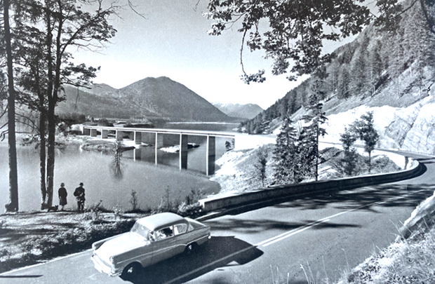 Schwarz-weiß-Foto mit einem Auto aus den 50er-Jahren im Vordergrund, das über eine geschwungene Straße fährt. Die Straße führt an einem See vorbei und als Brücke mit sieben Pfeilern über den See. Im Hintergrund sind Berge zu sehen.