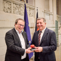 Der Verkehrsminister von Nordrhein-Westfalen übergibt symbolisch eine rote Fahrradklingel mit Aufschrift "VMK-Vorsitz" an Bayerns Verkehrsminister Christian Bernreiter.